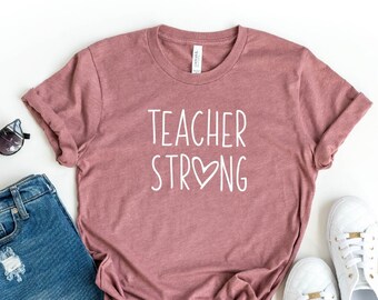May include: Heather mauve t-shirt with the text "Teacher Strong" in white with a heart symbol. The shirt is laid flat on a white background with a pair of sunglasses, denim shorts, and white sneakers.