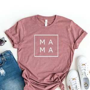 May include: A heather mauve t-shirt with the word "MAMA" in a white box on the front. The shirt is paired with denim shorts and white sneakers.