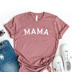 May include: A heather mauve t-shirt with the word "MAMA" printed in white on the front. The t-shirt is paired with denim shorts and white sneakers.