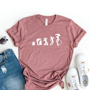 May include: A heather mauve t-shirt with a white graphic of the evolution of humans ending with an alien. The shirt is laid flat on a white background with a pair of sunglasses, denim shorts, and white sneakers.