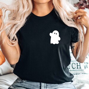 May include: A black t-shirt with a white ghost graphic on the front. The shirt is being worn by a person with long blonde hair. The person is sitting on a couch with a pillow featuring a pumpkin design.