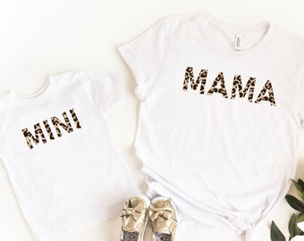 May include: Two white t-shirts, one adult and one child size, with the words "MAMA" and "MINI" in a leopard print design. Gold baby shoes and a bouquet of flowers are also in the image.
