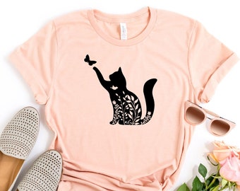 May include: A heather peach t-shirt with a black silhouette of a cat with flowers and a butterfly. The shirt is laid flat on a white background with a pair of grey shoes, sunglasses, and pink roses.