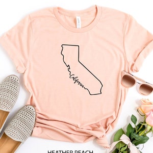 May include: A peach colored t-shirt with a black outline of the state of California and the word "California" written in cursive inside the outline. The shirt is laid flat on a white surface with a pair of beige shoes, sunglasses, and a bouquet of pink roses.