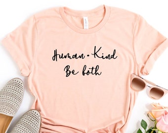 May include: A peach-colored t-shirt with the text "Human. Kind. Be both" in black script. The shirt is laid flat on a white background with a pair of gray shoes, sunglasses, and a bouquet of pink roses.
