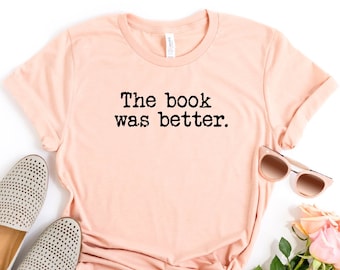 May include: A peach-colored t-shirt with the text "The book was better." printed in black. The shirt is laid flat on a white background with a pair of gray shoes, sunglasses, and a bouquet of pink roses.