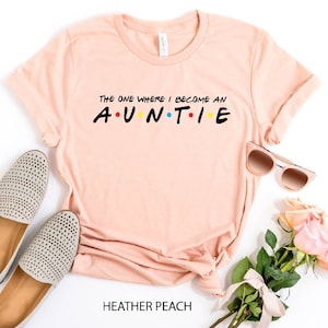 May include: A peach-colored t-shirt with the text "THE ONE WHERE I BECOME AN AUNTIE" in black with colorful dots between the letters.