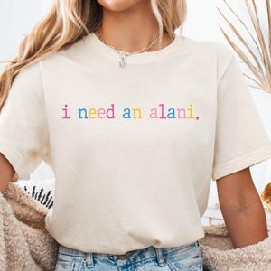 May include: A cream-colored t-shirt with the text "i need an alani" in rainbow colors. The shirt is paired with denim shorts. The word "Natural" is printed at the bottom left of the image.