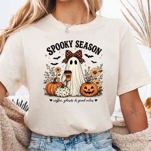May include: A cream-colored short-sleeve t-shirt featuring a graphic of a ghost wearing a plaid bow, holding a coffee cup, surrounded by pumpkins, sunflowers, and bats. The text reads SPOOKY SEASON and coffee, ghosts & good vibes.
