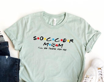 May include: A light blue t-shirt with the text "Soccer Mom I'll be there for you" in a black font. The text is styled to look like the "Friends" logo. The shirt is displayed on a white background with a pair of blue jeans and white trainers.