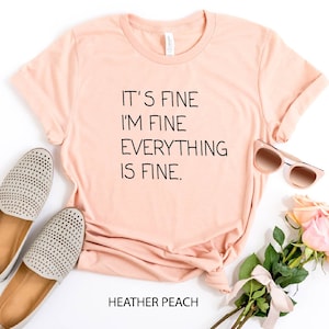 May include: A peach-colored t-shirt with the text "It's fine I'm fine Everything is fine." printed in black. The shirt is laid flat on a white surface with a pair of gray shoes, sunglasses, and pink roses.