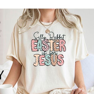 Może przedstawiać: Biała koszulka z napisem "Silly Rabbit Easter is for Jesus" w kolorowych literach z kropkami. Koszulka jest na modelce siedzącej na białym kocu.