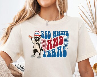 May include: Cream-colored t-shirt featuring a raccoon wearing a patriotic hat and holding a hot dog. The shirt has the text "RED WHITE AND TERRY" in red, white, and blue. The shirt is paired with denim shorts.