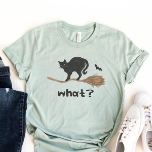 May include: Heather dusty blue t-shirt featuring a black cat riding a broom with the word "what?" below. The shirt is paired with dark blue jeans and white sneakers. A fun, casual Halloween-themed outfit.