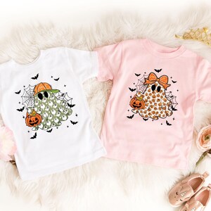Könnte beinhalten: Zwei kurzärmelige T-Shirts zum Thema Halloween. Eines ist weiß mit einem Geist, der eine Sonnenbrille und einen Kürbis trägt. Das andere ist rosa mit einem Geist, der eine Schleife trägt und einen Kürbis hält. Beide Shirts haben Spinnweben und Fledermäuse.