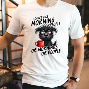 May include: White t-shirt with the text "I DON'T LIKE MORNING PEOPLE OR MORNINGS OR PEOPLE." The graphic features a black dog with red-rimmed glasses holding a red mug. The shirt is being worn by a person.