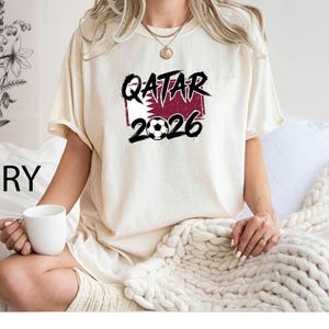 May include: Ivory-colored t-shirt with the text "QATAR 2026" in black, with a soccer ball graphic. The text is set against a maroon and white background. The shirt is short-sleeved.