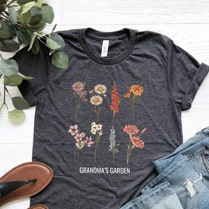 May include: Heather dark grey t-shirt featuring a floral print and the text "Grandma's Garden". The shirt is styled with blue jeans and black sandals.