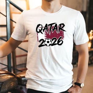 Może przedstawiać: Biała koszulka z napisem "QATAR 2026" w kolorze czarnym i grafiką piłki nożnej. Koszulka zawiera r&oacute;wnież flagę Kataru w bordowo-białym wzorze. Koszulka jest z kr&oacute;tkim rękawem.