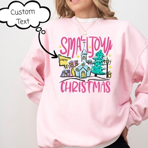 May include: A light pink sweatshirt with a colorful graphic that reads "Small Town Christmas." The graphic features a church, houses, a Christmas tree, and streetlights. The sweatshirt is a crew neck style.