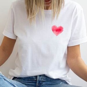May include: A white t-shirt with a red watercolor heart design on the front. The shirt is worn by a person sitting with their legs crossed, wearing blue jeans and white sneakers.