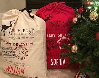 May include: Two red and white Santa sacks with the words "North Pole Post Office" and "Overnight Delivery" printed on them. The sacks are decorated with red and white Christmas stamps. The sacks are sitting on a beige tiled floor in front of a lit Christmas tree. The sacks are personalized with the names "William" and "Sophia".