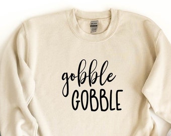 Op de afbeelding: Een zandkleurig sweatshirt met de tekst "gobble GOBBLE" in zwarte letters. Het sweatshirt wordt gecombineerd met een blauwe jeans en witte sneakers.