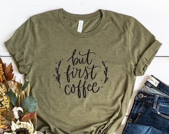 Puede incluir: Camiseta verde oliva con la frase "but first coffee" en negro. La imagen incluye vaqueros azules, botines abiertos color beige y un ramo de flores otoñales. La camiseta está anudada a la cintura.