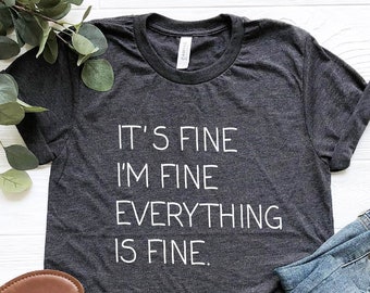 Può includere: Maglietta grigio scuro melange con il testo "It's fine I'm fine Everything is fine." La maglietta è mostrata su una superficie bianca con un paio di sandali marroni e un paio di jeans blu.