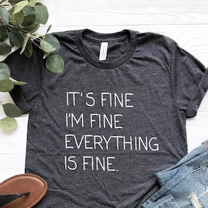 May include: Heather dark grey t-shirt with the text "It's fine I'm fine Everything is fine."  The shirt is shown on a white surface with a pair of brown sandals and blue jeans.