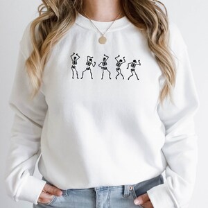 K&ouml;nnte beinhalten: Wei&szlig;er Pullover mit einem Motiv von f&uuml;nf tanzenden Skeletten. Die Skelette sind schwarz und schwarz umrandet.