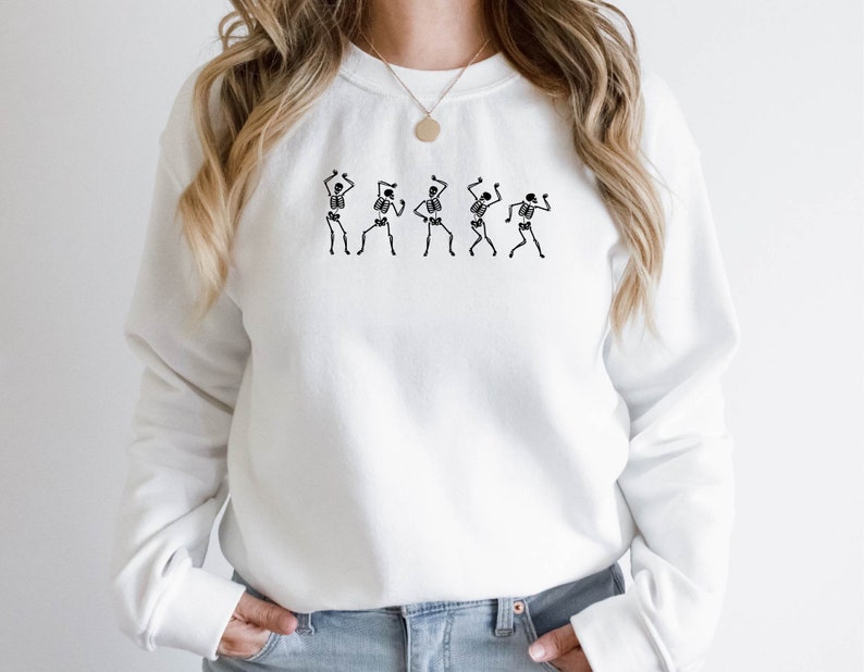 K&ouml;nnte beinhalten: Wei&szlig;er Pullover mit einem Motiv von f&uuml;nf tanzenden Skeletten. Die Skelette sind schwarz und schwarz umrandet.
