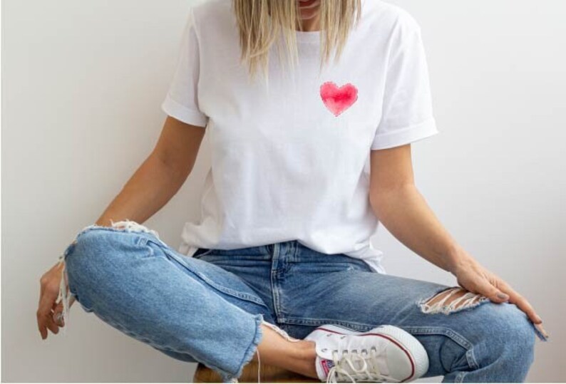 May include: A white t-shirt with a red watercolor heart design on the front. The shirt is worn by a person sitting with their legs crossed, wearing blue jeans and white sneakers.