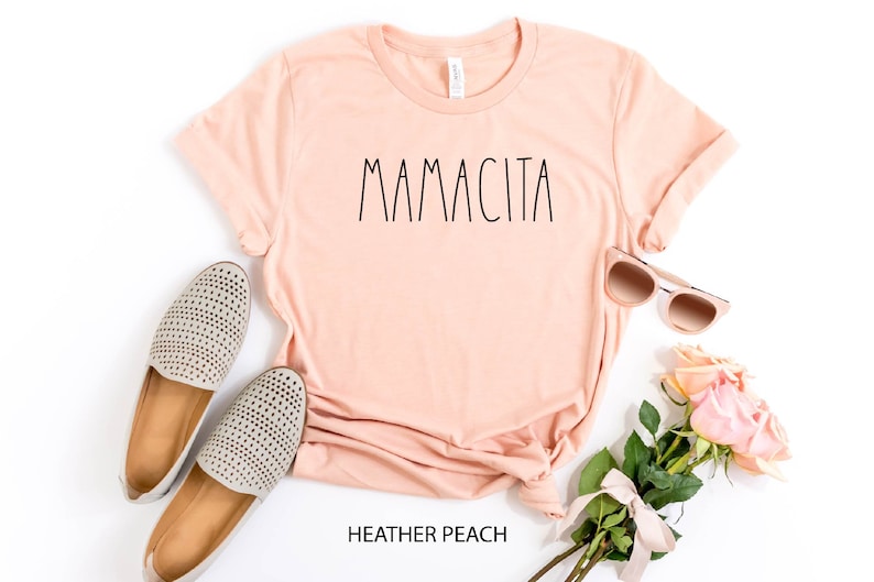 Może przedstawiać: R&oacute;żowy t-shirt z napisem "MAMACITA" w czarnych literach. Koszulka leży płasko na białym tle z parą szarych but&oacute;w, okularami przeciwsłonecznymi i bukietem r&oacute;żowych r&oacute;ż.