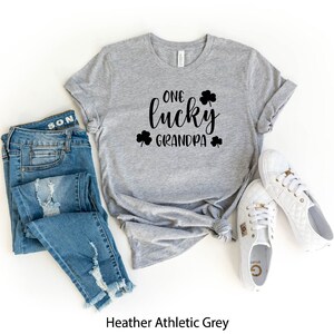 Puede incluir: Camiseta gris deportivo heather con el texto "One lucky grandpa" en letras negras y dise&ntilde;os de tr&eacute;boles. La camiseta se combina con jeans azules y zapatillas blancas.