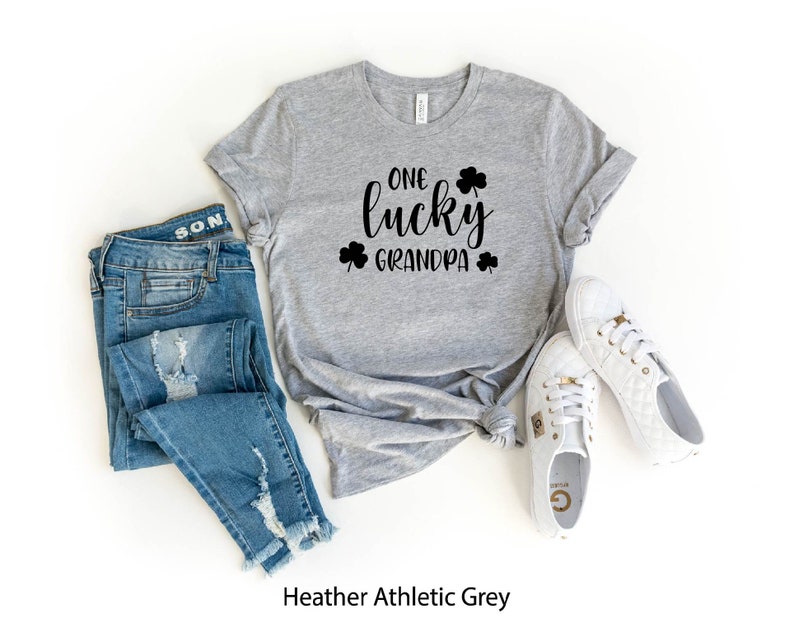 Puede incluir: Camiseta gris deportivo heather con el texto "One lucky grandpa" en letras negras y dise&ntilde;os de tr&eacute;boles. La camiseta se combina con jeans azules y zapatillas blancas.