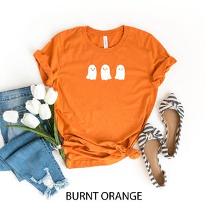 K&ouml;nnte beinhalten: Orangefarbenes T-Shirt mit drei wei&szlig;en Geister-Silhouetten auf der Vorderseite. Das Shirt liegt flach auf einem wei&szlig;en Hintergrund mit einem Paar blau-wei&szlig; gestreiften Schuhen und einer blauen Jeans mit wei&szlig;en Blumen.