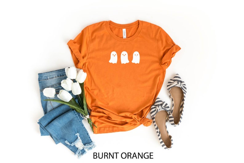 K&ouml;nnte beinhalten: Orangefarbenes T-Shirt mit drei wei&szlig;en Geister-Silhouetten auf der Vorderseite. Das Shirt liegt flach auf einem wei&szlig;en Hintergrund mit einem Paar blau-wei&szlig; gestreiften Schuhen und einer blauen Jeans mit wei&szlig;en Blumen.