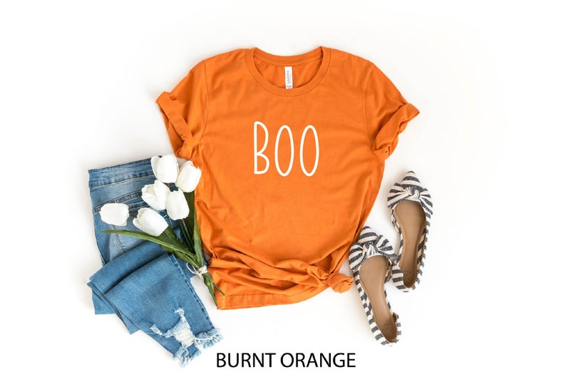 May include: Burnt orange t-shirt with the word "BOO" in white, paired with blue jeans, white tulips, and striped flats. The shirt is folded, and the jeans are distressed. The flats have a bow detail.