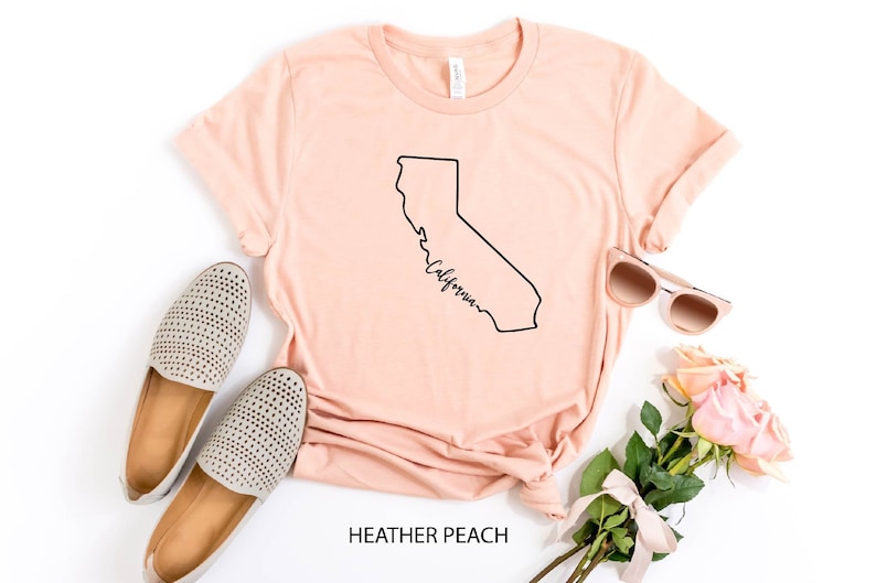 May include: A peach colored t-shirt with a black outline of the state of California and the word "California" written in cursive inside the outline. The shirt is laid flat on a white surface with a pair of beige shoes, sunglasses, and a bouquet of pink roses.