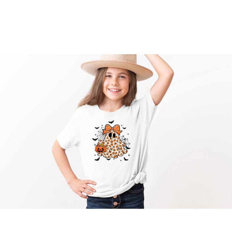 May include: White t-shirt featuring a Halloween ghost design. The ghost is adorned with a pumpkin pattern, an orange bow, and a jack-o'-lantern. Black bats and spiderwebs complete the festive design, perfect for the spooky season.