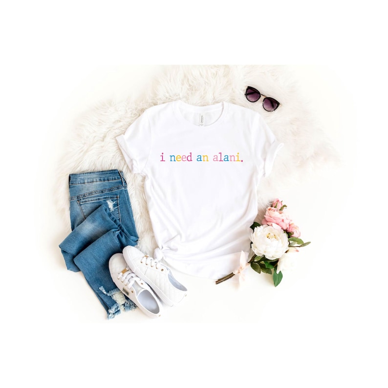 May include: White t-shirt with the text "i need an alani" in rainbow colors. The shirt is styled with blue jeans, white sneakers, sunglasses, and a bouquet of pink and white flowers on a white fluffy surface.