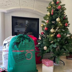 May include: Two green and red Santa sacks with a reindeer graphic and the text "North Pole Post Office" and "Overnight Delivery". The sacks are sitting in front of a fireplace with a decorated Christmas tree.