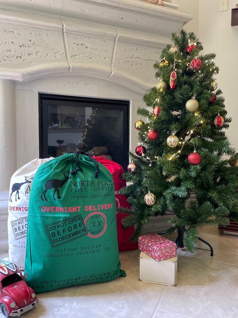 May include: Two green and red Santa sacks with a reindeer graphic and the text "North Pole Post Office" and "Overnight Delivery". The sacks are sitting in front of a fireplace with a decorated Christmas tree.