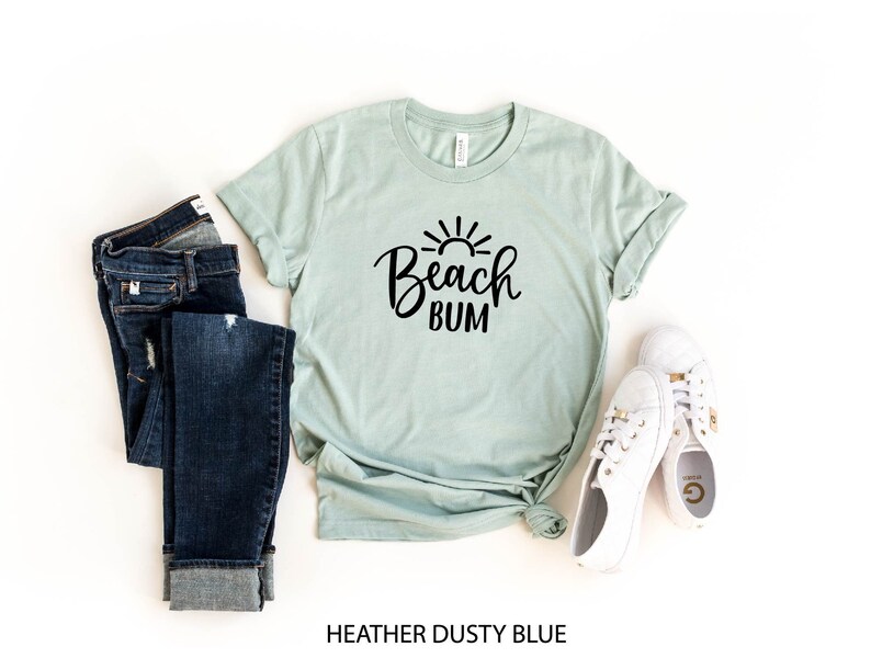 Puede incluir: Una camiseta azul claro con un gr&aacute;fico negro que dice "Beach Bum" y un gr&aacute;fico de sol sobre el texto. La camiseta est&aacute; colocada sobre un fondo blanco con un par de jeans azules y zapatillas blancas.