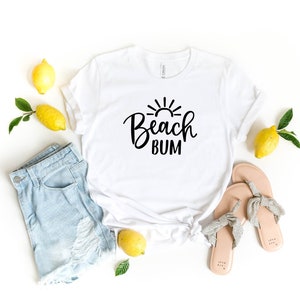 Puede incluir: Camiseta blanca con texto negro que dice "Beach Bum" y un gr&aacute;fico de sol sobre el texto. La camiseta est&aacute; colocada sobre una superficie blanca con limones, unos pantalones cortos de mezclilla y un par de sandalias.