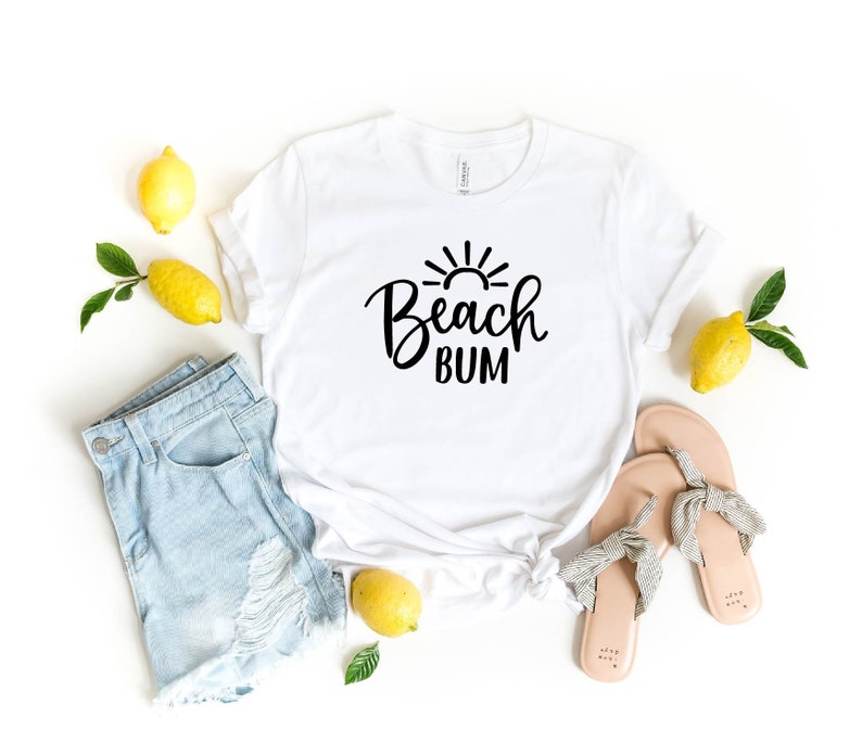Puede incluir: Camiseta blanca con texto negro que dice "Beach Bum" y un gr&aacute;fico de sol sobre el texto. La camiseta est&aacute; colocada sobre una superficie blanca con limones, unos pantalones cortos de mezclilla y un par de sandalias.
