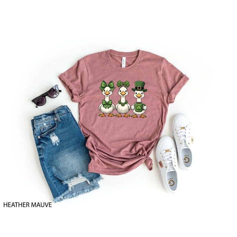 May include: Heather mauve t-shirt with a St. Patrick's Day design featuring three cartoon geese wearing green bows, shamrocks, and a leprechaun hat. The shirt is paired with denim shorts, sunglasses, and white sneakers.
