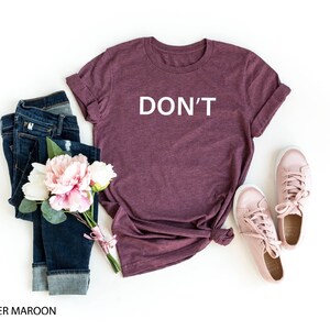Pu&ograve; includere: Una t-shirt color marrone rossiccio con la scritta "DON'T" stampata in bianco sul davanti. La t-shirt &egrave; stesa su uno sfondo bianco con un paio di scarpe da ginnastica rosa e un paio di jeans blu.