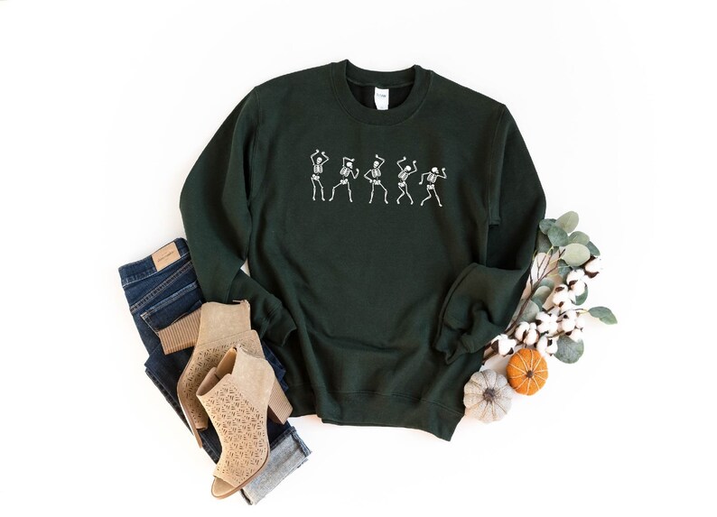 K&ouml;nnte beinhalten: Ein dunkelgr&uuml;ner Pullover mit einer wei&szlig;en Grafik von f&uuml;nf tanzenden Skeletten. Der Pullover liegt flach auf einem wei&szlig;en Hintergrund mit einem Paar braunen Stiefeletten, blauen Jeans und einem Zweig Eukalyptus mit Baumwollkn&auml;ueln.